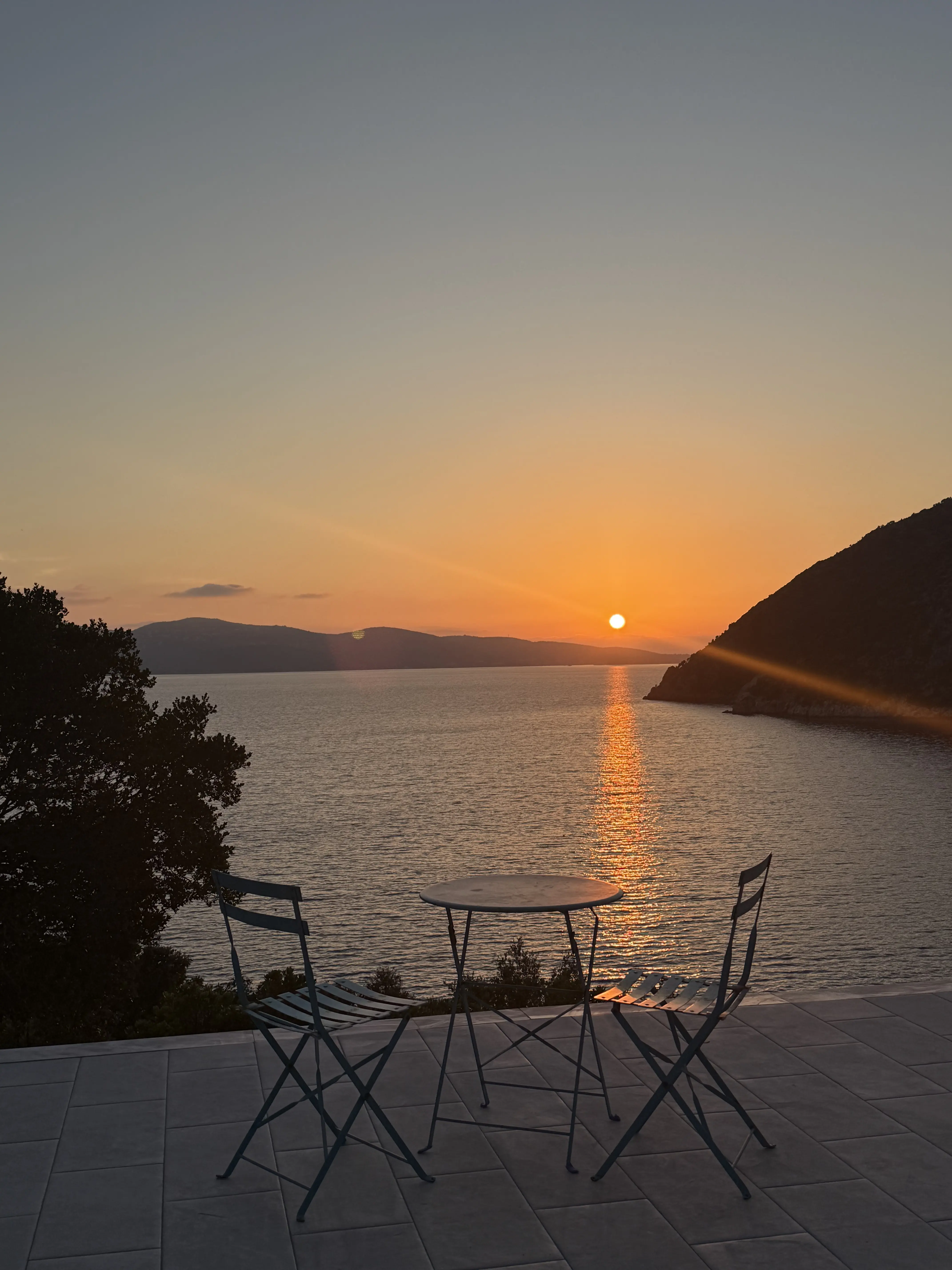 A small table and two chairs on the terrace at sunset, looking west over the water toward Kefalonia